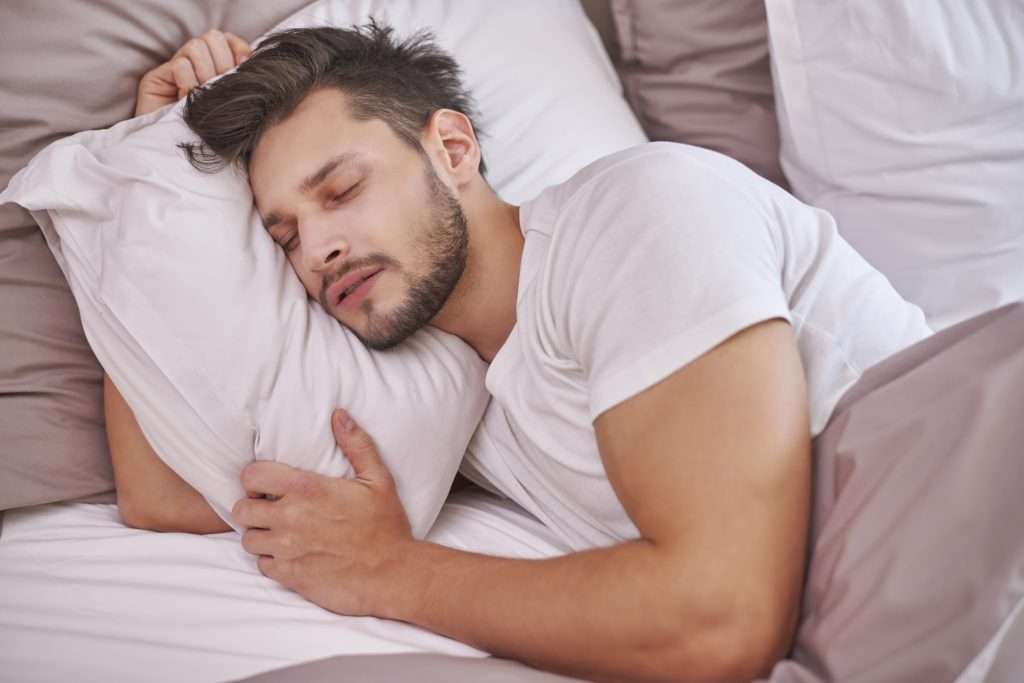 homem de barba e camiseta branca dormindo abraçado ao travesseiro