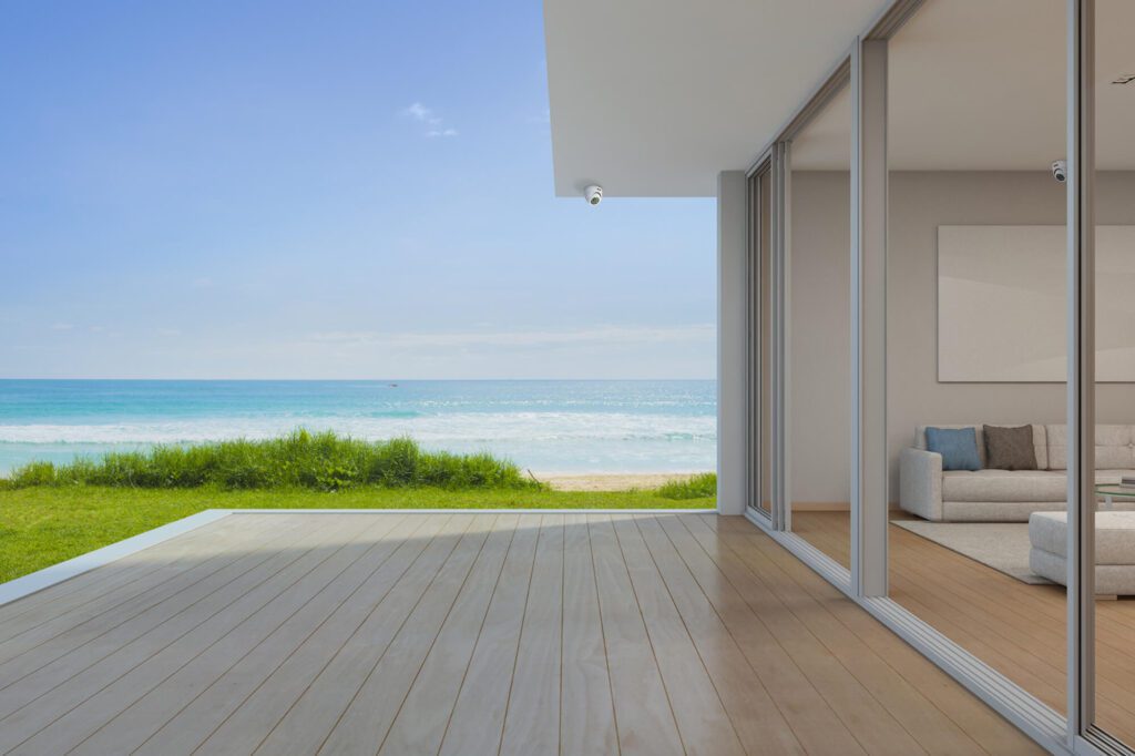 foto em perspectiva de área em casa de praia com vidraças tendo ao fundo restinga e o mar azul claro