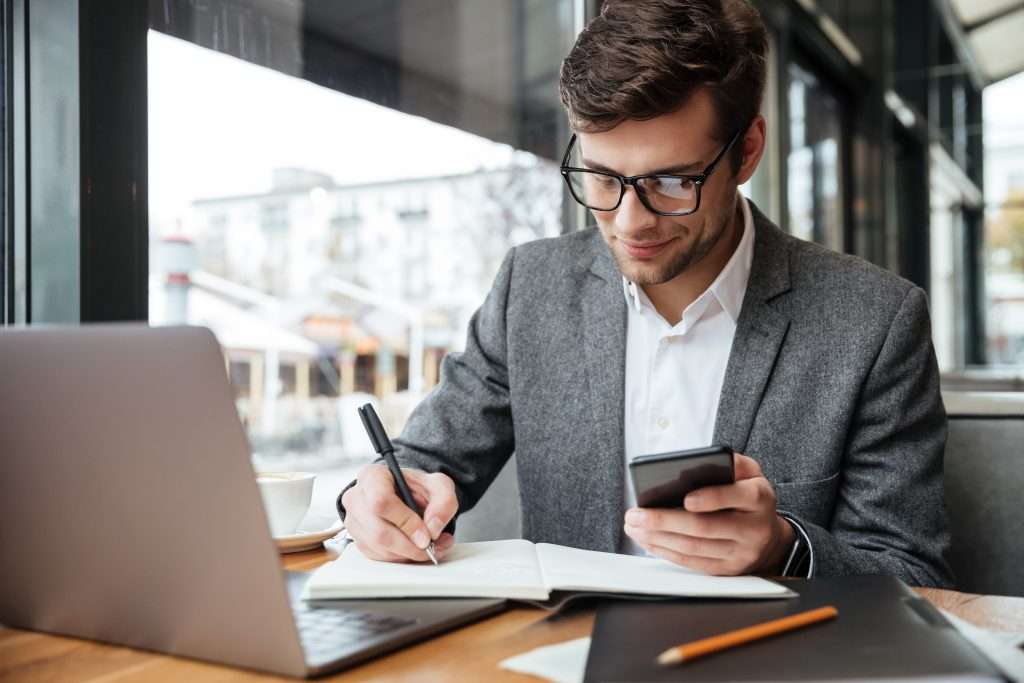 homem de terno com de celular sentado na frente de notebook escrevendo documento