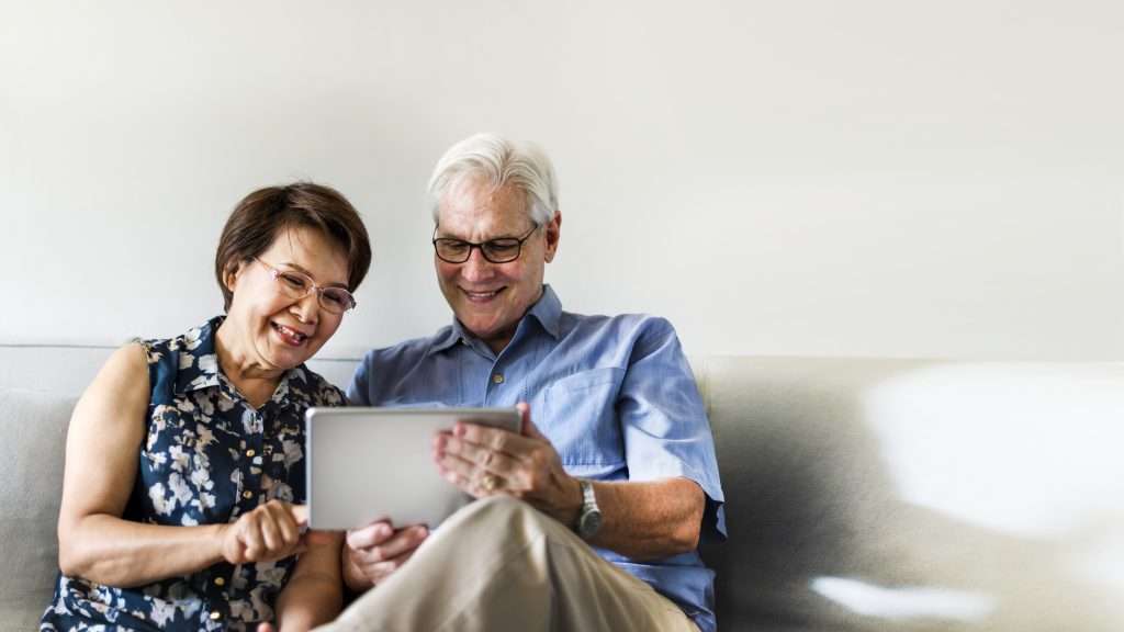 casal de idosos sorrindo sentados no sofá lendo notícia em um tablet