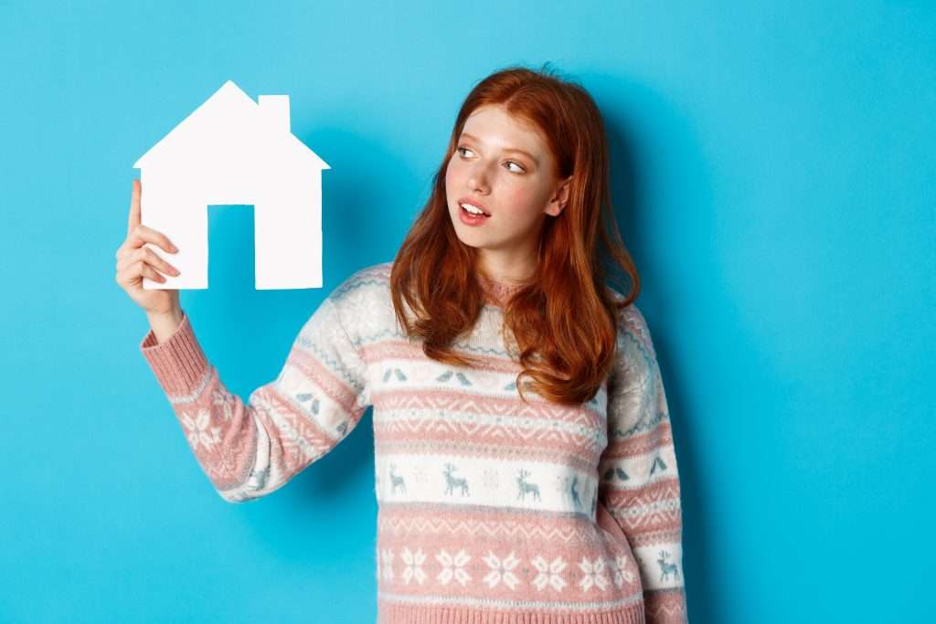 Mulher de cabelos ruivos trajando blusa de lã estampada segurando miniatura de casa branca em uma das mãos em fundo azul claro