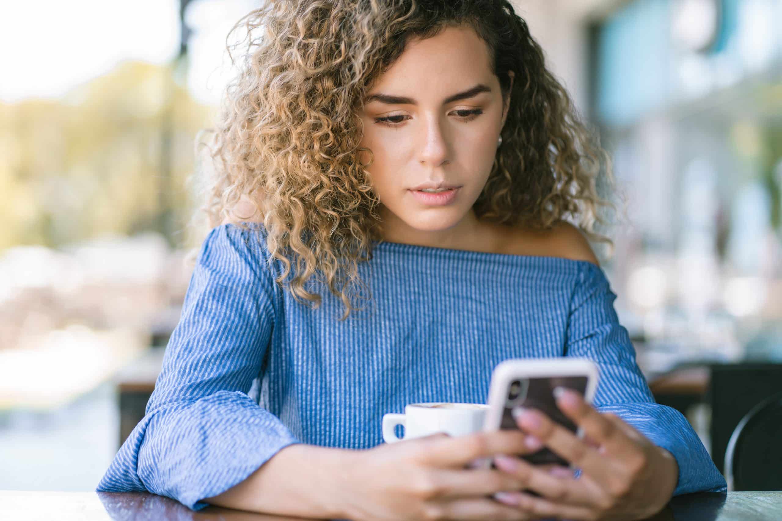 mulher de cabelo crespo com blusa azul clara lendo no celular com fundo desfocado