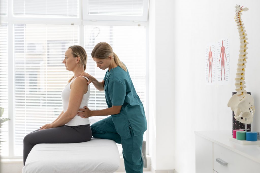 fisioterapeuta com uniforme verde musgo em seu consultório pressionando mãos nas costas de cliente idosa de camiseta de alças brancas sentada na beirada da cama