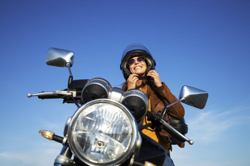 Mulher feliz trajando jaqueta marrom com óculos escuros sentada em motocicleta ajustando capacete com look de motociclista e céu azul ao fundo